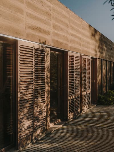 Architectural project - wooden shutters, earth wall, neutral vibes, residential exterior, earthy finish, environment blending, natural materials, wood, timber, outdoor space, natural light, shadow play, warm tones, neutral tones, beige, brown, exterior design, low carbon design, daylight optimization, eco friendly, rustic modern, organic design, minimalist design, rammed earth, rammed earth wall, green architecture, earthy texture, modern rustic, sustainable home, passive house, natural ventilation, landscape integration, clean lines, timber cladding, modern architecture, energy efficient, earthy tones, inviting exterior, rustic exterior, sustainable facade, warm exterior, contemporary facade, brown tones, tactile surfaces, eco facade
