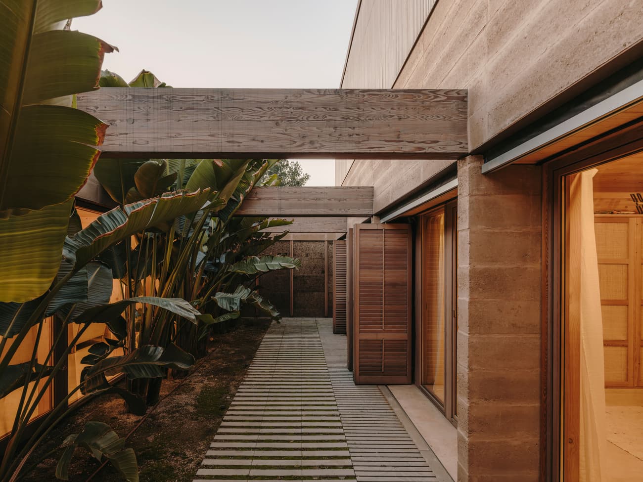 Architectural project - wooden shutters, outdoor corridor, calm space, tropical vibe, tropical exterior, concrete walls, open spaces, tropical plants, exterior living, nature integrated, natural materials, serene environment, natural light, sustainable design, warm tones, innovative design, contemporary style, green, daylight, beige, light wood, eco friendly, wooden beams, timber beams, natural textures, minimalist design, tranquil setting, rammed earth, low carbon, natural palette, passive house, modern facade, modern architecture, earthy materials, earthy tones, warm atmosphere, cozy outdoor, residential design, garden view, inviting exterior, lush greenery, warm lighting, architectural harmony, indoor outdoor flow, seamless design