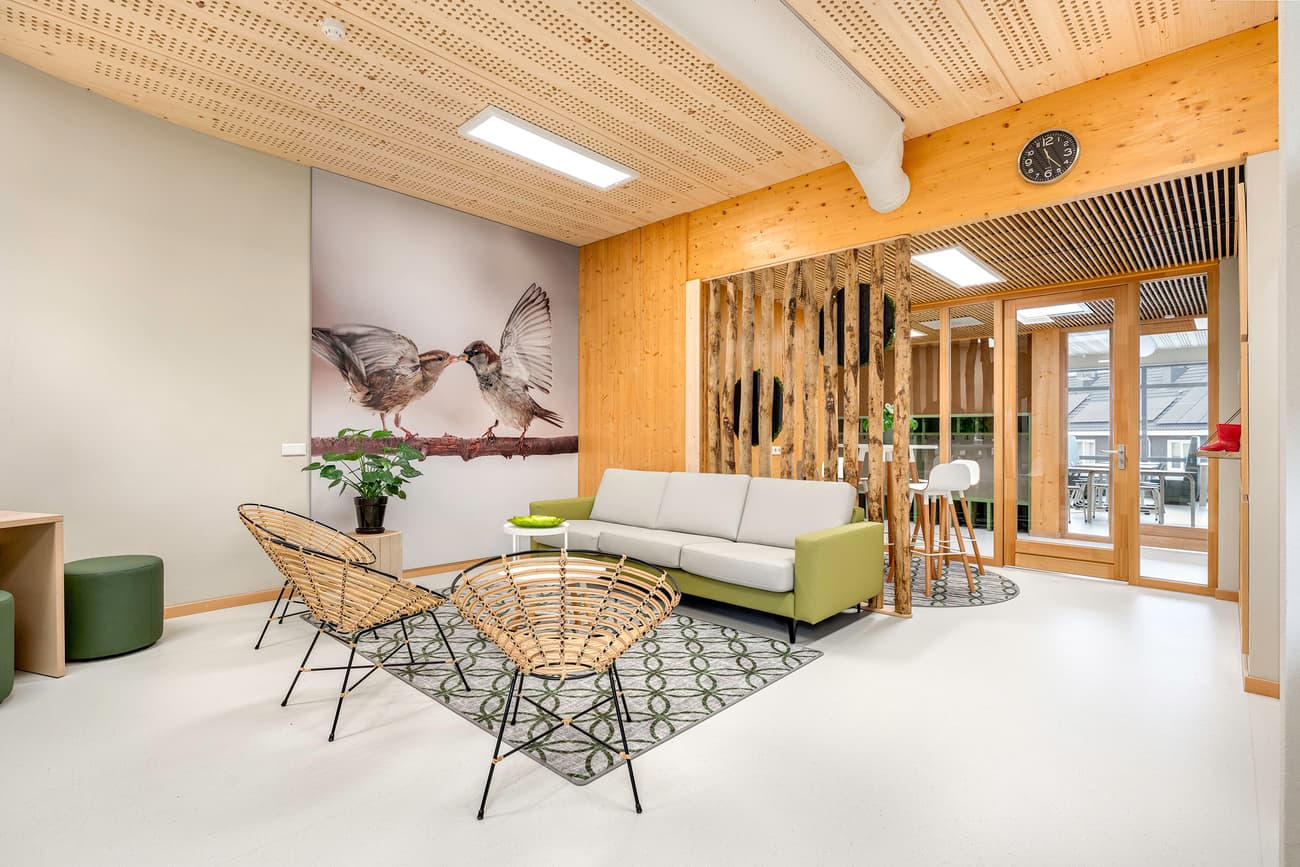 Architectural project - green accents, open ceiling, sustainable space, relaxed environment, cozy lounge, office lounge, break area, modern office, collaborative area, informal meeting, networking space, nature theme, bird mural, large mural, decorative wall, soft seating, modern chairs, rattan chairs, patterned rug, natural materials, soft lighting, wood, timber, fabric, natural light, inviting atmosphere, sustainable design, glass, warm tones, eco design, green, neutral tones, daylight, wooden ceiling, beige, light wood, multifunctional space, eco friendly, honey wood, wood beams, Scandinavian style, casual seating, glass doors, circular design, white, Nordic design, timber interior, open plan, greenery, seating area, comfortable seating, bright space, timber wall, indoor plants, wood interior, geometric rug