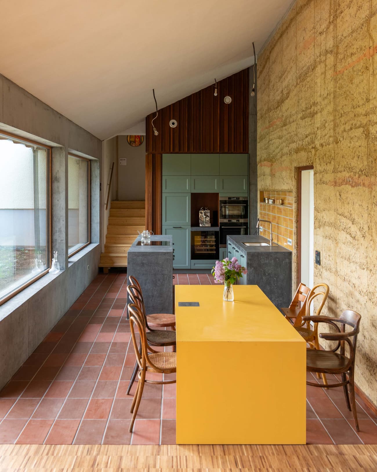 Architectural project - cozy dining, wood chair, muted palette, farmhouse table, vintage chairs, indoor space, eco-friendly design, wood, timber, steel, natural light, yellow, warm tones, grey, daylight, beige, concrete, low carbon design, rustic vibe, architectural lighting, Scandinavian style, wooden floor, rammed earth wall, earth tones, modern rustic, sustainable home, passive house, large windows, glass window, home kitchen, open layout, dining table, inviting space, natural texture, terracotta, warm vibe, modern kitchen, kitchen island, compact kitchen, sunlit room, open space, functional kitchen, terracotta floor