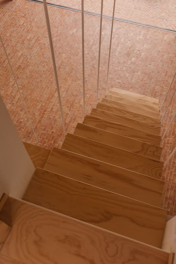 Architectural project - vibrant floor, herringbone pattern, upcycled materials, Nordic touch, minimalist railing, white railing, brick floor, warm space, wood stairs, vertical railing, home staircase, indoor stairs, DIY style, sustainable flooring, recycled brick, natural materials, architectural detail, interior view, wood, timber, natural light, warm tones, contemporary style, daylight, beige, light wood, functional design, modern design, Scandinavian influence, compact space, honey wood, minimalist style, cozy space, eco-friendly materials, clean lines, simple elegance, inviting space, handcrafted look, terracotta, brick, cozy atmosphere, residential design, warm vibe, earthy textures, staircase view, terracotta floor, timber steps, wooden stair, red