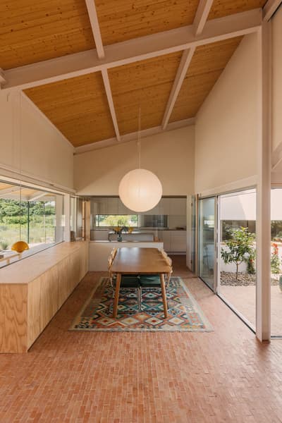 Architectural project - family dining, home dining, cozy dining, modern farmhouse, Scandinavian cabin, simple table, pattern rug, large pendant, brick tiles, kitchen view, pink, wood, timber, natural light, sustainable design, pastel colors, glass, warm tones, neutral tones, exposed beams, daylight, daylight optimization, honey wood, natural textures, Scandinavian style, glass doors, multifunctional, modern rustic, eco home, passive house, white walls, indoor outdoor, white, inviting home, Nordic style, timber ceiling, rustic style, comfortable home, relaxed living, casual living, open layout, open plan, dining area, dining table, inviting space, earthy tones, brick, garden view, warm vibe, wooden elements, bright space, residential, home, wood ceiling, airy atmosphere, playful space, indoor, renovation, terracotta floor, minimalist shelves, colorful rug, remodel