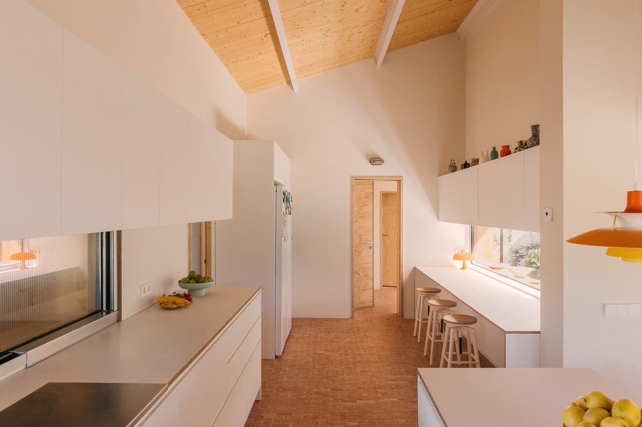 Architectural project - breakfast bar, pendant lights, brick floor, cream, white cabinets, sleek cabinets, colorful decor, playful kitchen, modern stools, Nordic feel, inviting kitchen, kitchen nook, pastel decor, cozy interior, natural materials, wood, timber, natural light, sustainable design, warm tones, daylight, wooden ceiling, light wood, daylight optimization, eco friendly, Scandinavian influence, honey wood, neutral palette, clean lines, white, timber ceiling, family kitchen, home kitchen, bright kitchen, casual dining, terracotta, residential space, minimalist kitchen, modern kitchen, compact kitchen, efficient layout, warm lighting, open shelving, terracotta floor, sloped ceiling