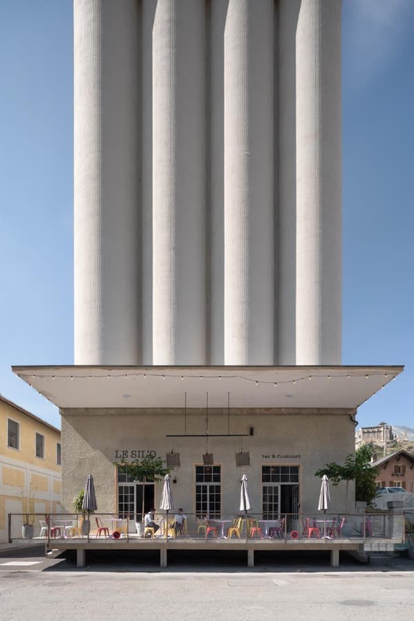 Architectural project - raw concrete, urban renewal, modern renovation, urban chic, city center, rooftop terrace, concrete facade, architectural landmark, historic building, historic preservation, architectural heritage, heritage site, colorful furniture, concrete columns, industrial architecture, brutalist architecture, cafe exterior, vibrant chairs, cafe umbrellas, towering structure, open air dining, mix of old and new, contrasting elements, industrial revival, street cafe, urban setting, metal, cityscape, grey, industrial vibe, beige, concrete, neutral palette, minimalist design, outdoor cafe, public space, outdoor seating, adaptive reuse, urban design, modern facade, white, textured facade, architectural contrast