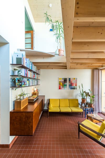 Architectural project - playful interior, mezzanine, terracotta tiles, potted plants, cozy living space, red tile floor, yellow sofa, retro sofa, colorful art, vibrant art, architect design, wood, timber, natural light, sustainable design, yellow, warm tones, neutral tones, exposed beams, ceiling beams, daylight, eco friendly, timber beams, natural textures, Scandinavian style, relaxed atmosphere, contemporary home, clean lines, white, Nordic style, casual living, open layout, inviting space, minimalist decor, terracotta, warm vibe, wooden elements, modern living room, reading nook, home library, bright interior, earthy textures, wood ceiling, family room, wooden furniture, open shelving, ceramic, floating shelves, spacious feel, indoor plants, mid-century modern, double height