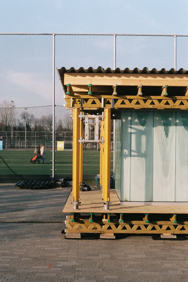 Architectural project - temporary structure, minimal design, timber, urban setting, quick assembly, grey, metal, timber frame, passive design, yellow, glass, modular construction, prefabricated building, modular design, eco design, sustainable construction, green building, yellow framework, exposed structure, corrugated roof, transparent walls, community space, outdoor area, sports facility, athletic site, simple structure, industrial aesthetic, contemporary architecture, public use, urban landscape, versatile structure, innovative design, lightweight construction, flexible design, space-saving design, outdoor enclosure, engineered timber, contemporary style, modern vibe, urban park, city environment, sustainable architecture, renewable materials, dynamic space, multifunctional area, assembly design, green, neutral tones