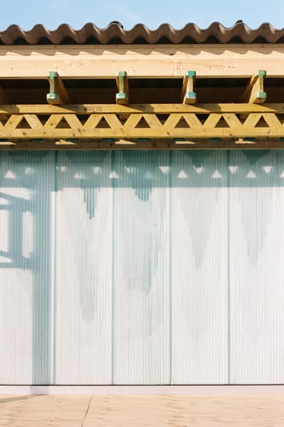 Architectural project - light blue, natural materials, architectural detail, eco-friendly design, wood, timber, raw materials, steel, metal, outdoor space, shadow play, natural tones, corrugated roof, contemporary architecture, sustainable architecture, neutral tones, modern construction, daylight, beige, wood facade, timber facade, truss structure, exposed trusses, light and shadow, wavy roof, architectural elements, structural design, roof design, roof detail, timber detail, wooden trusses, timber trusses, exterior design, building facade, architectural facade, minimal aesthetics, minimalist facade, geometric pattern, visual rhythm, light wood, natural wood, exterior wall, open air design, modern aesthetic, architectural photography, building design, golden