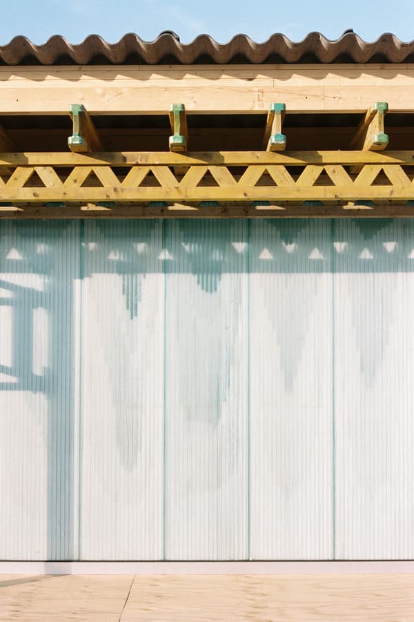 Architectural project - light blue, natural materials, architectural detail, eco-friendly design, wood, timber, raw materials, steel, metal, outdoor space, shadow play, natural tones, corrugated roof, contemporary architecture, sustainable architecture, neutral tones, modern construction, daylight, beige, wood facade, timber facade, truss structure, exposed trusses, light and shadow, wavy roof, architectural elements, structural design, roof design, roof detail, timber detail, wooden trusses, timber trusses, exterior design, building facade, architectural facade, minimal aesthetics, minimalist facade, geometric pattern, visual rhythm, light wood, natural wood, exterior wall, open air design, modern aesthetic, architectural photography, building design, golden
