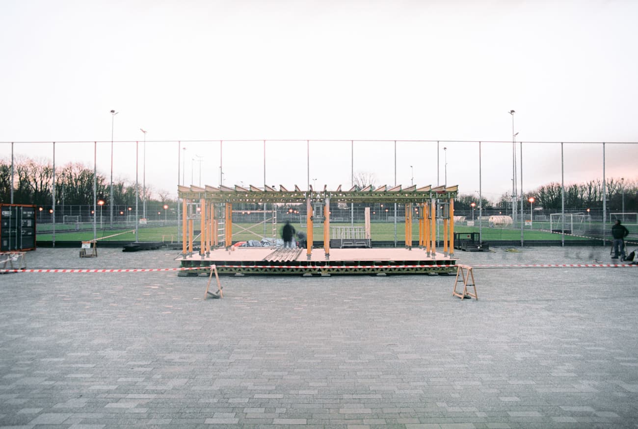 Architectural project - wood, timber, temporary structure, urban setting, grey, metal, wooden frame, sustainable design, modular design, flexible design, urban park, neutral tones, public installation, urban environment, industrial vibe, sports venue, minimalist construction, multifunctional space, eco friendly, contemporary design, open framework, outdoor pavilion, public space, gathering area, metal mesh, community center, outdoor seating, event space, architectural assembly, field backdrop, adaptable structure, lightweight materials, social hub, outdoor venue, modern gathering, light construction, streamlined architecture