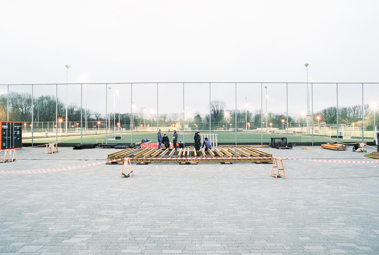 Architectural project - temporary structure, wood, timber, urban setting, grey, outdoor space, natural light, community space, neutral tones, construction site, construction workers, outdoor construction, urban development, construction process, sports field, building site, open area, gathering space, fencing, safety barriers, timber planks, wood framework, evening light, spacious layout, public area, athletic grounds, city park, pavement, concrete paving, large scale, structural work, outdoor setting, sports venue, recreational area, public project, civic space, landscape architecture, field lights, modern infrastructure, urban planning, community development, wide shot, neighborhood park, evening scene, cold climate, beige, brown, concrete
