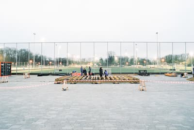 Architectural project - temporary structure, wood, timber, urban setting, grey, outdoor space, natural light, community space, neutral tones, construction site, construction workers, outdoor construction, urban development, construction process, sports field, building site, open area, gathering space, fencing, safety barriers, timber planks, wood framework, evening light, spacious layout, public area, athletic grounds, city park, pavement, concrete paving, large scale, structural work, outdoor setting, sports venue, recreational area, public project, civic space, landscape architecture, field lights, modern infrastructure, urban planning, community development, wide shot, neighborhood park, evening scene, cold climate, beige, brown, concrete