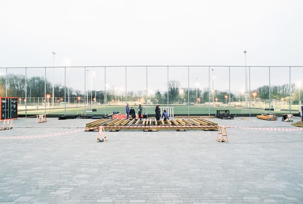 Architectural project - temporary structure, wood, timber, urban setting, grey, outdoor space, natural light, community space, neutral tones, construction site, construction workers, outdoor construction, urban development, construction process, sports field, building site, open area, gathering space, fencing, safety barriers, timber planks, wood framework, evening light, spacious layout, public area, athletic grounds, city park, pavement, concrete paving, large scale, structural work, outdoor setting, sports venue, recreational area, public project, civic space, landscape architecture, field lights, modern infrastructure, urban planning, community development, wide shot, neighborhood park, evening scene, cold climate, beige, brown, concrete