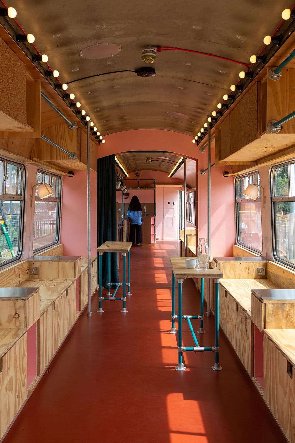 Architectural project - bright red, red floor, train interior, pink walls, train carriage, mobile kitchen, pop-up restaurant, street food, terracotta flooring, wooden countertops, metal table, retro style, vintage touch, quirky design, playful interior, unique restaurant, portable venue, travel inspired, brightly lit, railway car, adventurous dining, steel legs, pastel pink, pink, wood, metal, natural light, sustainable design, recycled materials, plastic, warm tones, modular design, industrial vibe, beige, functional design, blue, eco friendly, compact space, flexible use, vibrant space, creative space, modern rustic, open plan, social gathering, casual dining, cozy atmosphere, interior, pine wood, warm lighting, colorful space, urban style, red