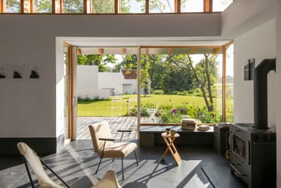 Architectural project - minimal design, cozy interior, eco-friendly design, natural light, inviting atmosphere, passive design, eco-conscious, daylight, multifunctional space, Scandinavian style, neutral palette, light tones, modern rustic, sustainable home, large windows, indoor outdoor, landscape view, wood accents, nature-inspired, relaxing space, outdoor connection, rural setting, garden view, nature immersion, comfortable seating, minimalist aesthetic, bright interior, glass door, open living, lounge area, wooden frames, timber frames, Nordic vibes, warm living, sunlit room, fluid transition, modern cottage, simple decor, black floor, slate floor, garden seating, compact living, efficient layout, small table, modern stove, hearth, indoor warmth