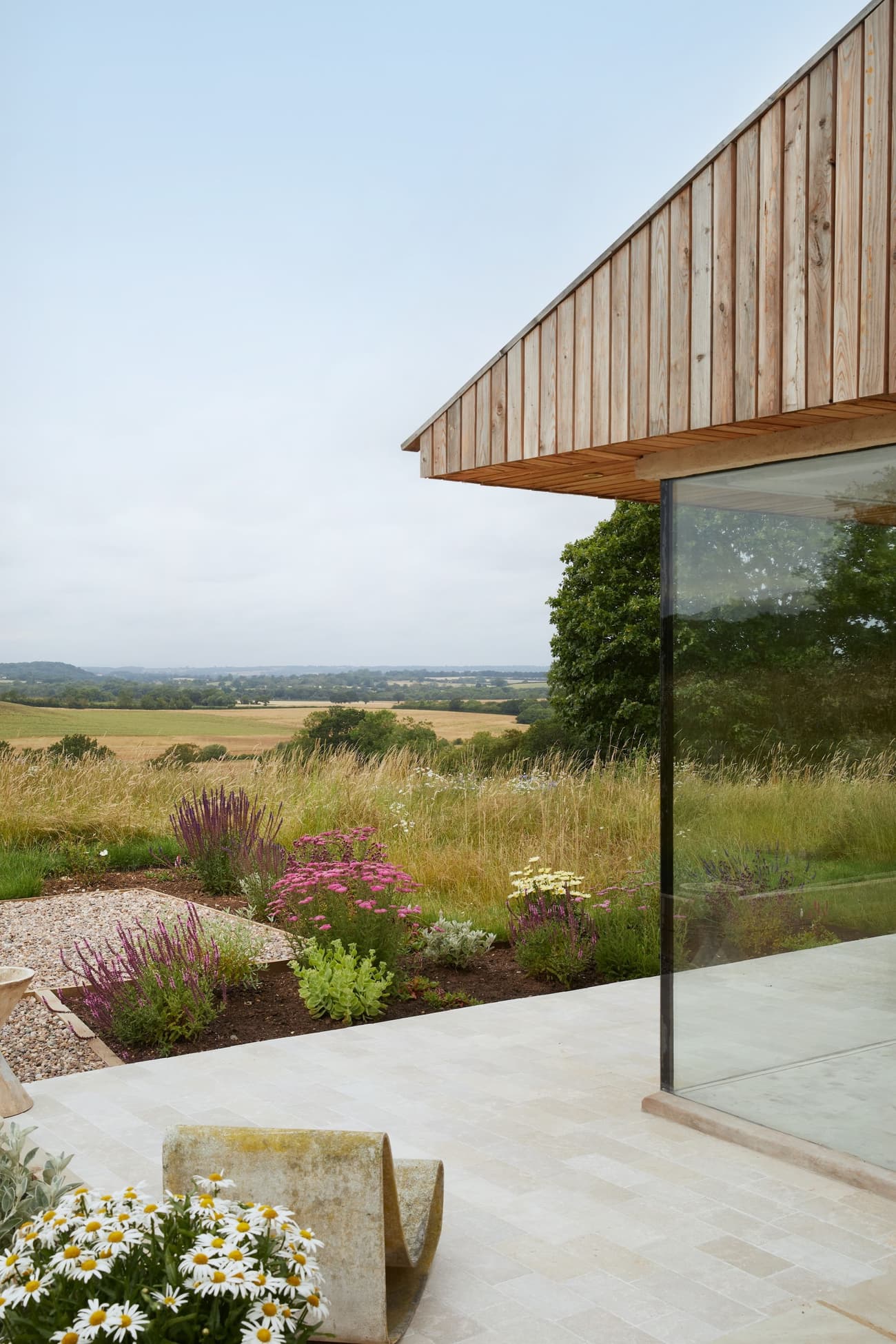 Architectural project - timber, passive design, sustainable design, glass, eco design, neutral tones, beige, wood facade, low carbon design, natural textures, minimalist design, outdoor seating, blue sky, light tones, natural landscape, biophilic design, scenic view, clean lines, timber cladding, wood cladding, countryside home, outdoor view, stone, panoramic view, greenery, nature integration, serene setting, eco house, patio area, seamless integration, peaceful atmosphere, glass wall, rural house, rural retreat, modern retreat, modern house, terrace garden, open horizon, glazed corner, wildflowers, natural garden, minimal look, tranquility, natural beauty