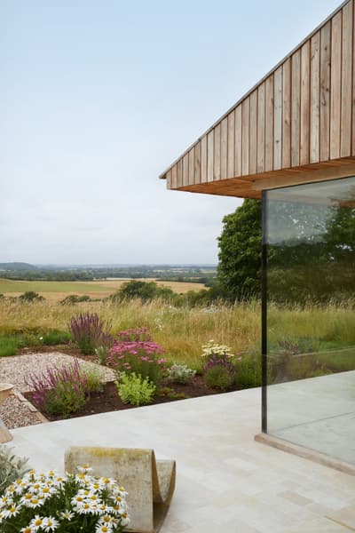 Architectural project - timber, passive design, sustainable design, glass, eco design, neutral tones, beige, wood facade, low carbon design, natural textures, minimalist design, outdoor seating, blue sky, light tones, natural landscape, biophilic design, scenic view, clean lines, timber cladding, wood cladding, countryside home, outdoor view, stone, panoramic view, greenery, nature integration, serene setting, eco house, patio area, seamless integration, peaceful atmosphere, glass wall, rural house, rural retreat, modern retreat, modern house, terrace garden, open horizon, glazed corner, wildflowers, natural garden, minimal look, tranquility, natural beauty