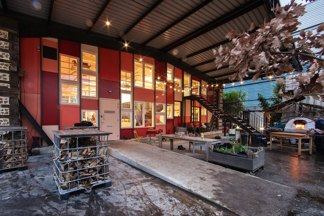Architectural project - exterior facade, red panels, industrial facade, modern industrial, urban renewal, steel staircase, vibrant atmosphere, stacked wood, terrace area, city space, vibrant red, urban outdoor, social place, workshop style, urban terrace, metal elements, public gathering, collaborative space, lively setting, wood, steel, metal, outdoor space, rustic touch, sustainable design, recycled materials, glass, exposed structure, corrugated roof, community space, city environment, grey, beige, concrete, industrial style, multifunctional space, eco friendly, community hub, outdoor seating, urban design, creative space, workshop area, large windows, open layout, concrete floor, warm lighting, glass facade, daylight use, wood storage, exterior space, urban vibe, red, metal ceiling