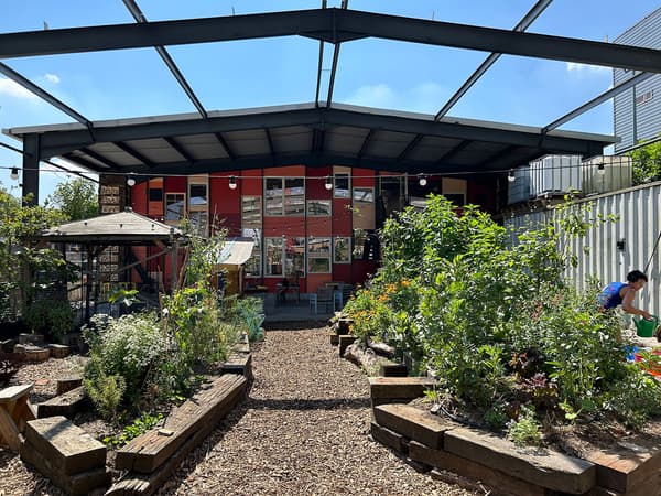 Architectural project - garden venue, green oasis, red wall, vibrant color, raised planters, native plants, public venue, city escape, casual vibes, wooden benches, air circulation, active space, colorful facade, urban farming, community garden, garden beds, relaxation space, natural materials, wood, steel, metal, passive design, sustainable design, glass, warm tones, eco design, community space, outdoor area, green, neutral tones, daylight, industrial vibe, recreational area, beige, brown, industrial elements, eco friendly, sunlight, leisure space, open concept, social space, gathering area, outdoor seating, steel structure, modern rustic, biophilic design, metal roof, inviting space, environmental design, nature retreat, tranquil atmosphere, open air, glass windows, lush greenery, educational space, urban garden, red
