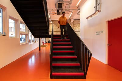 Architectural project - metallic finishes, contrasting colors, industrial staircase, metal stairs, steel staircase, red steps, industrial interior, exposed ductwork, exposed pipes, warehouse vibe, loft style, commercial space, meeting room, orange floor, vivid floor, bold colors, vibrant atmosphere, mixed-use space, overhead lighting, raw aesthetic, collaborative environment, innovation hub, urban office, ventilation system, eco-friendly design, steel, metal, utilitarian design, green building, neutral tones, functional space, concrete, modern design, architectural lighting, minimalist design, black, adaptive reuse, bright colors, orange, industrial chic, creative space, workspace, white walls, white, open plan, black railing, open space, sustainable renovation, high ceiling, urban style, red