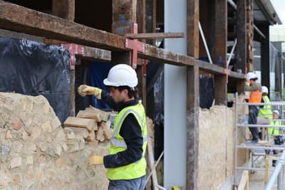 Architectural project - natural stone, renovation process, building renovation, historical restoration, brick wall, stone masonry, protective gear, safety helmet, high-visibility vest, industrial work, traditional techniques, restoration project, unfinished wall, construction worker, stone laying, masonry work, architecture in progress, brick laying, structural repair, wall repair, material reuse, work site safety, outdoor work, building expertise, skilled labor, construction craftsmanship, architectural conservation, heritage preservation, natural materials, wood, scaffolding, raw materials, timber frame, sustainable construction, construction site, site work, urban development, construction materials, building site, beige, brown, wooden beams, stone, brick, stone wall, heritage building, craftsmanship, eco renovation