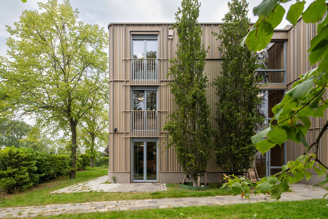 Architectural project - vertical elements, green facade, modern residence, tree-lined, eco-friendly design, wood, timber, outdoor space, eco-conscious, contemporary style, neutral tones, landscape architecture, light wood, natural setting, exterior, glass doors, eco building, green architecture, biophilic design, passive house, natural ventilation, modern facade, timber cladding, harmonious design, energy efficient, green living, nature-inspired, environmental architecture, nature integration, wood slats, wooden facade, residential building, sustainable facade, energy saving, urban nature, vertical cladding, wood architecture, architectural integration, sustainable living, apartment building