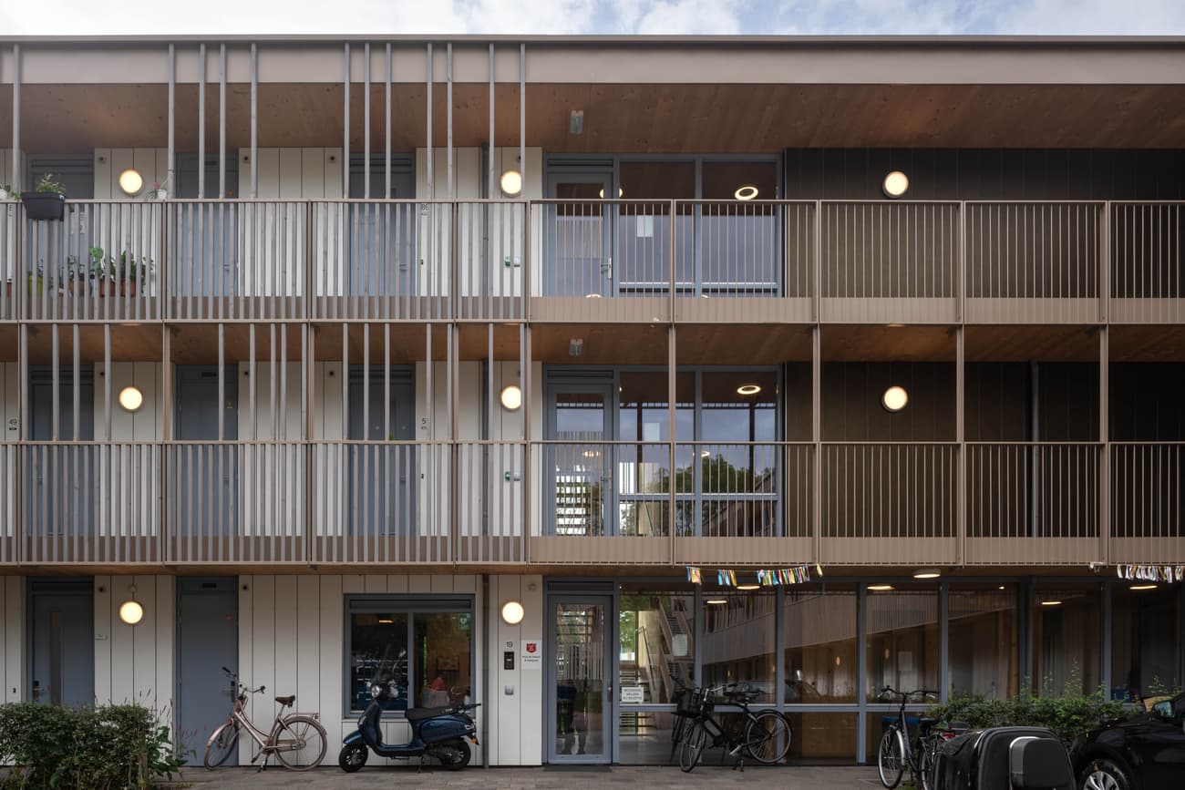 Architectural project - apartment facade, sustainable housing, metal railing, multi-story building, wooden details, warm facade, simple geometry, community living, eco-friendly architecture, bicycle friendly, shared spaces, natural materials, wood, timber, urban lifestyle, metal, glass, warm tones, innovative design, contemporary style, neutral tones, urban development, beige, brown, wood facade, timber facade, exterior design, light wood, daylight optimization, Scandinavian influence, neutral palette, minimalist design, passive house, natural ventilation, modern architecture, energy efficient, green living, residential building, balcony design, functional layout, modern living, vertical slats, architectural rhythm, urban housing