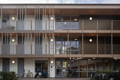 Architectural project - apartment facade, sustainable housing, metal railing, multi-story building, wooden details, warm facade, simple geometry, community living, eco-friendly architecture, bicycle friendly, shared spaces, natural materials, wood, timber, urban lifestyle, metal, glass, warm tones, innovative design, contemporary style, neutral tones, urban development, beige, brown, wood facade, timber facade, exterior design, light wood, daylight optimization, Scandinavian influence, neutral palette, minimalist design, passive house, natural ventilation, modern architecture, energy efficient, green living, residential building, balcony design, functional layout, modern living, vertical slats, architectural rhythm, urban housing