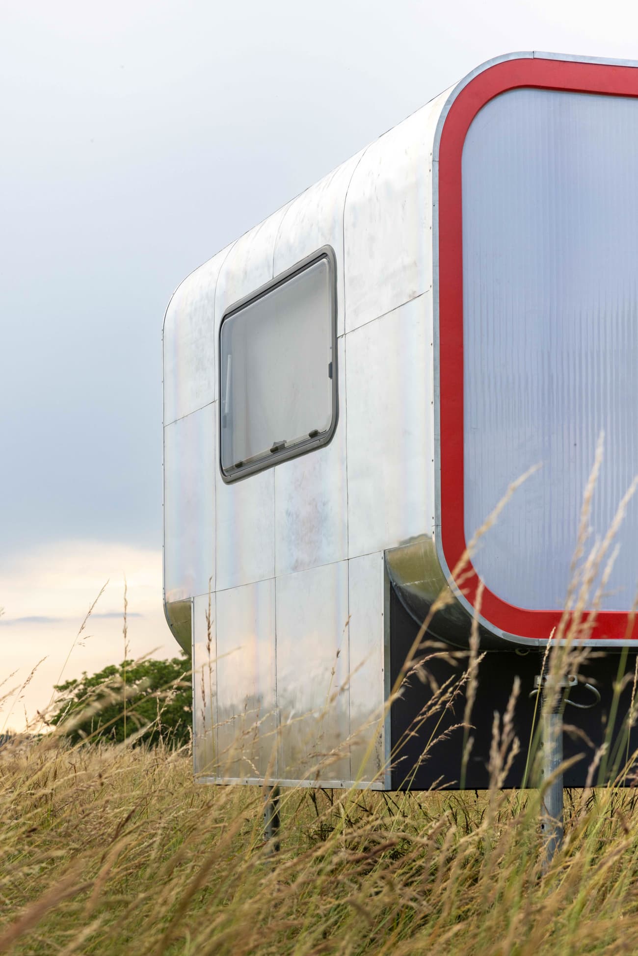 Architectural project - tiny house, flexible living, micro home, futuristic home, metal cladding, aluminum facade, curved corners, rounded edges, transportable home, contemporary dwelling, mobile residence, trailer home, field setting, silver exterior, red accent, sunlight reflection, eco living, modern caravan, inventive design, innovative dwelling, prefab home, prefabricated construction, outdoor placement, streamlined exterior, eco-friendly design, sustainable materials, grey, metal, glass, modular design, creative architecture, adaptable design, industrial style, modern design, natural setting, sleek lines, minimalist design, lightweight structure, mobile unit, open landscape, sustainable home, white, rural home, off-grid living, minimalist aesthetic, sleek design, compact living, aluminum, architectural innovation, red, modern materials