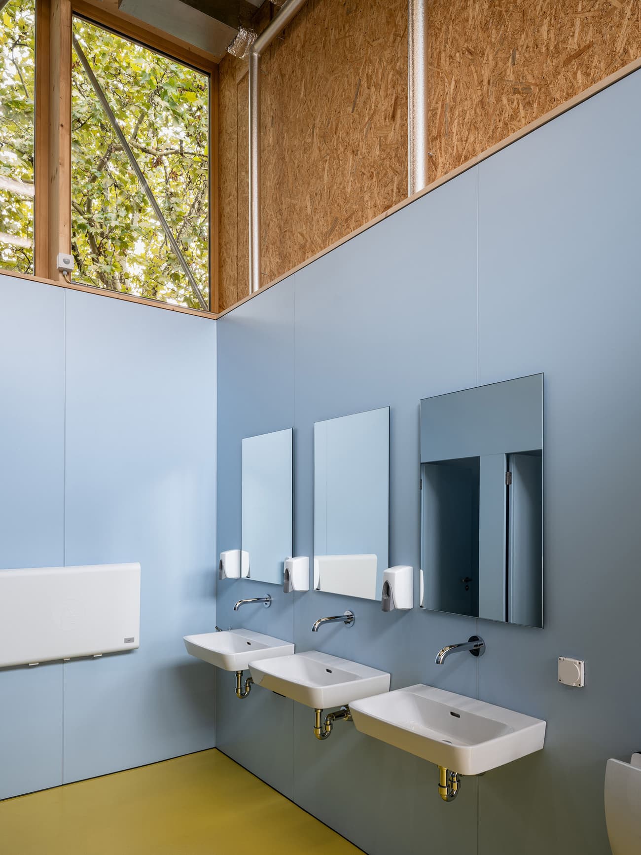 Architectural project - pastel blue, ventilation system, contemporary washroom, wall-mounted sinks, floating sinks, rectangular mirrors, hygiene station, public restroom, blue walls, OSB panels, sleek faucets, light yellow floor, yellow flooring, bathroom fixtures, restroom mirrors, light blue, eco-friendly design, timber, steel, natural light, yellow, glass, natural tones, eco design, sustainable architecture, daylight, plywood, beige, natural wood, low carbon design, blue, Scandinavian influence, compact space, neutral palette, minimalist design, clean aesthetic, wood panels, exposed wood, durable materials, clean lines, large windows, Nordic design, timber accents, bright interior, functional layout, practical space, soft tones, modern bathroom, urban style