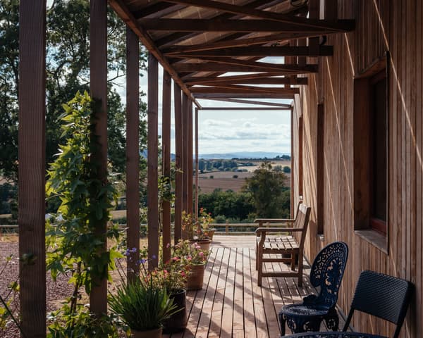 Architectural project - peaceful environment, potted plants, cozy patio, wooden pergola, shaded area, wooden siding, country style, black chairs, wood deck, timber terrace, plant pots, blue chairs, natural materials, wood, timber, outdoor space, inviting atmosphere, outdoor living, sustainable design, warm tones, natural tones, eco design, brown, light wood, blue, natural setting, rustic vibe, rustic charm, outdoor seating, tranquil setting, timber cladding, landscape view, countryside view, rural view, earthy tones, relaxing space, environmentally friendly, garden view, open air, wooden furniture, rural retreat, outdoor bench