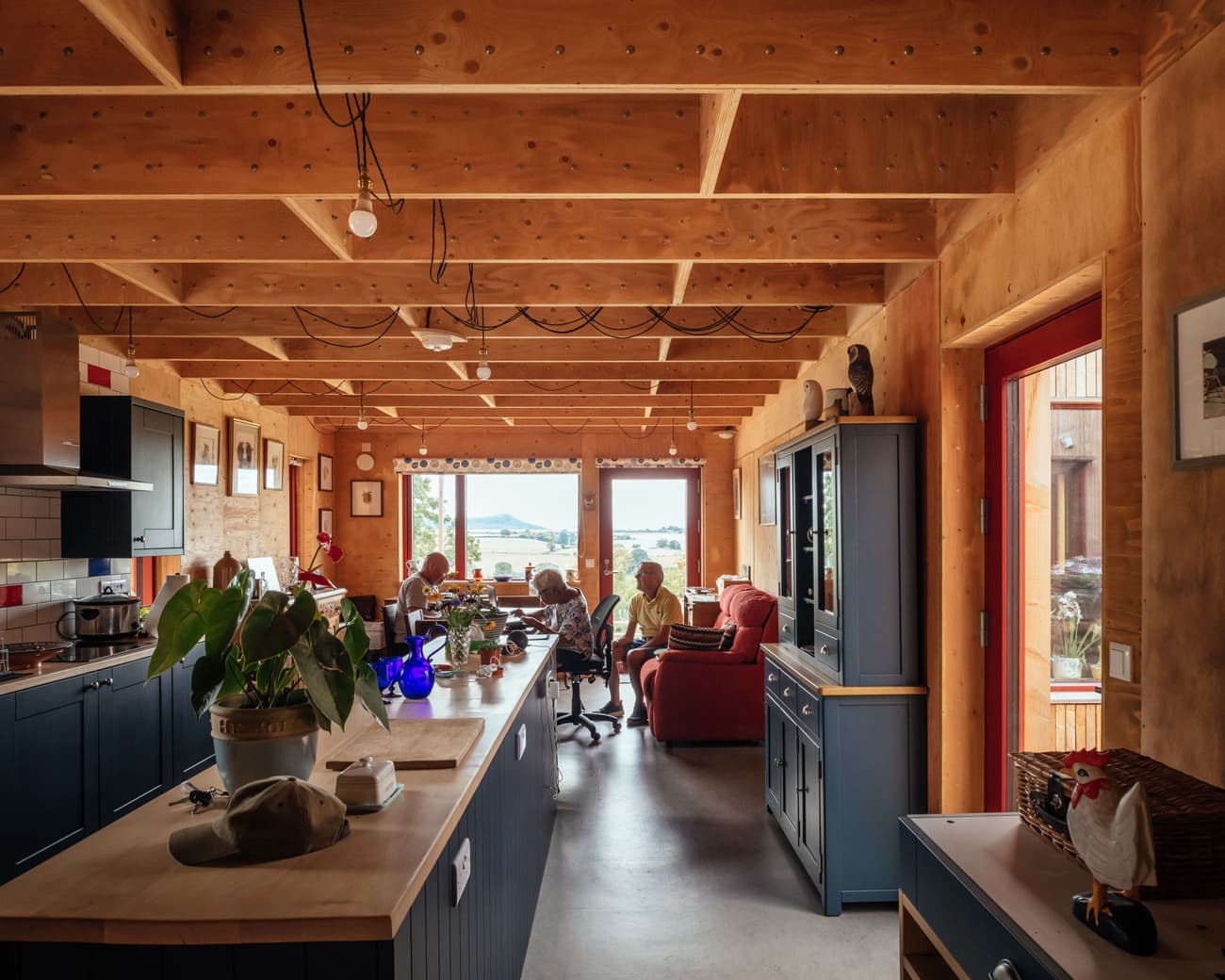Architectural project - cottage vibe, pendant lights, social area, ceiling lights, timber home, blue cabinetry, comfortable setting, blue kitchen, coastline home, cozy interior, natural materials, leisure area, wood, timber, natural light, inviting atmosphere, sustainable design, warm tones, natural tones, exposed structure, exposed beams, daylight, multi-use space, wooden ceiling, gathering space, beige, concrete, blue, wooden beams, Scandinavian style, casual seating, timber walls, modern rustic, nature view, eco friendly home, Nordic style, wood walls, relaxed living, wood cabin, open plan, living area, earthy tones, family gathering, rustic design, textured walls, rustic aesthetic, waterfront view, warm living, concrete floor, family room, wooden furniture, homey feel, indoor plants, red