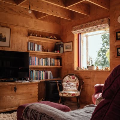 Architectural project - living room, countryside vibe, homey atmosphere, country style, soft seating, personal touches, floral chair, patterned chair, upholstered furniture, vintage chair, sofa set, throw blanket, decorative pillows, nature outside, inviting room, bookshelves, decor display, upholstery, intimate space, natural materials, wood, timber, relaxation area, natural light, warm tones, natural tones, neutral tones, exposed beams, beige, brown, light wood, sunlight, honey wood, rustic charm, timber walls, window view, wooden interior, wood walls, rural style, cozy corner, traditional style, raw finish, reading nook, home library, interior, home, built-in shelving, warm living room, cozy living room, residential interior, living space