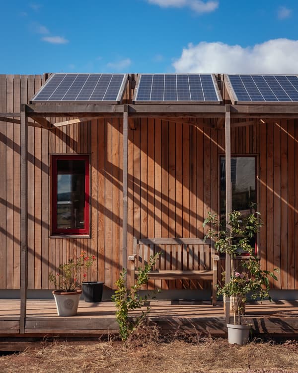 Architectural project - natural shade, warmth, solar panels, potted plants, contemporary barn, serene ambiance, plant pots, porch, sunny exterior, exterior sitting area, minimal design, natural materials, wooden bench, wood, timber, natural light, shadow play, sustainable design, warm tones, eco design, simple structure, brown, wood facade, timber facade, daylight optimization, outdoor leisure, natural textures, outdoor seating, raw wood, green architecture, modern rustic, eco home, cozy vibe, countryside home, energy efficient, rustic style, earthy tones, rural setting, inviting exterior, wooden deck, passive solar, vertical cladding, sustainable living