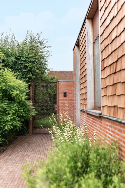 Architectural project - contemporary exterior, exterior walkway, modern landscape, warm facade, red bricks, terracotta cladding, outdoor path, brick path, rust, natural materials, sustainable materials, outdoor space, eco design, green, brown, exterior design, urban oasis, rustic charm, neutral colors, modern facade, earthy tones, terracotta, landscape design, brick facade, greenery, lush garden, private residence, clay tiles, exterior cladding, red, green plants