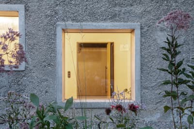 Architectural project - facade, sustainability, natural facade, light grey, residential exterior, concrete frame, natural landscaping, home exterior, square window, yellow interior, cozy light, inviting glow, outer wall, taupe, sustainable design, yellow, eco design, green, concrete, exterior wall, simple design, serene atmosphere, minimalist design, tranquil setting, low carbon, eco home, sustainable home, passive house, countryside home, outdoor view, textured wall, plaster wall, plaster, rough texture, nature-inspired, greenery, garden view, warm light, peaceful vibe, rustic exterior, modern exterior, window frame, wildflowers