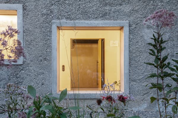 Architectural project - facade, sustainability, natural facade, light grey, residential exterior, concrete frame, natural landscaping, home exterior, square window, yellow interior, cozy light, inviting glow, outer wall, taupe, sustainable design, yellow, eco design, green, concrete, exterior wall, simple design, serene atmosphere, minimalist design, tranquil setting, low carbon, eco home, sustainable home, passive house, countryside home, outdoor view, textured wall, plaster wall, plaster, rough texture, nature-inspired, greenery, garden view, warm light, peaceful vibe, rustic exterior, modern exterior, window frame, wildflowers